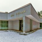 Gray concrete building with the sign 'Дом ученых' above the entrance, snow on the ground and large windows along the left side.