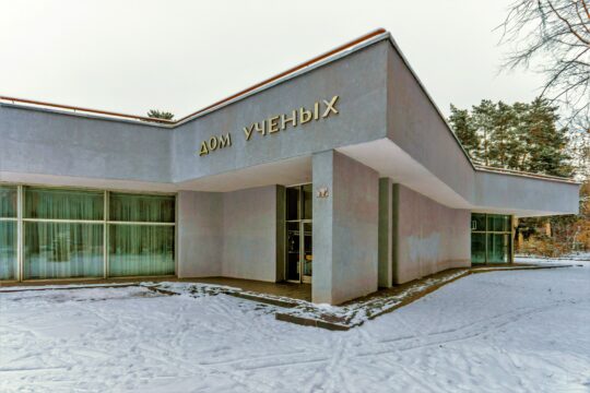 Gray concrete building with the sign 'Дом ученых' above the entrance, snow on the ground and large windows along the left side.